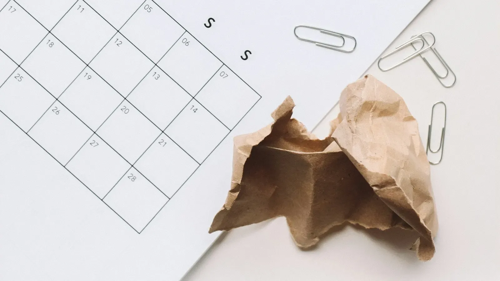 A minimalist printed monthly calendar grid on a white desk with paper clips and a crumpled brown paper bag