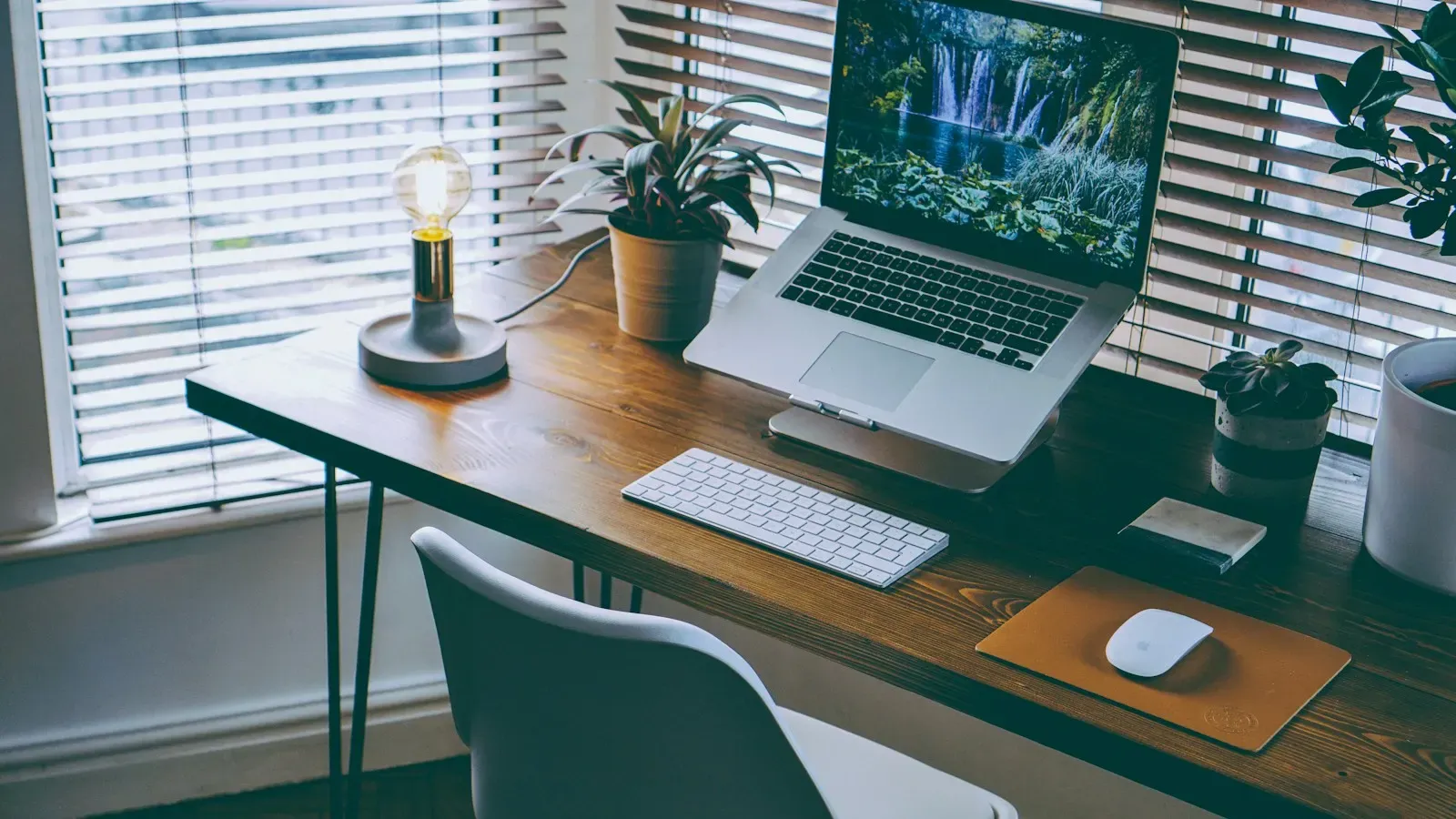 A laptop on a wooden desk by a window with blinds, with a desk lamp, a keyboard, a mouse and potted plants