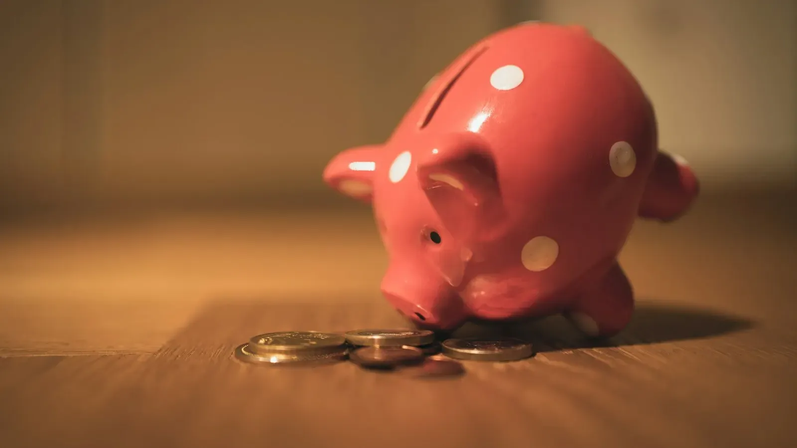A pink polka-dot piggy bank on a wooden table, nosing a small pile of coins