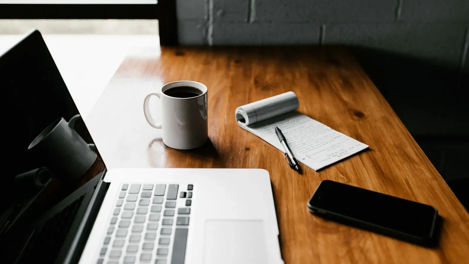 An open laptop on a wooden table next to a white coffee mug, a notepad with a pen, and a smartphone